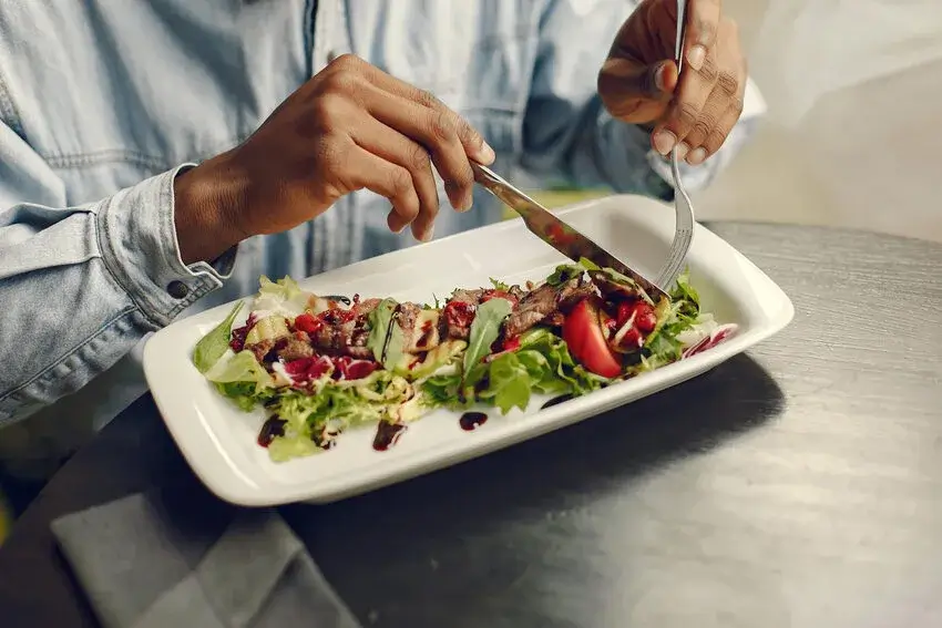 A person with dark skin is using a knife and fork to eat a salad with meat, vegetables, and a sauce sitting in vegan restaurant