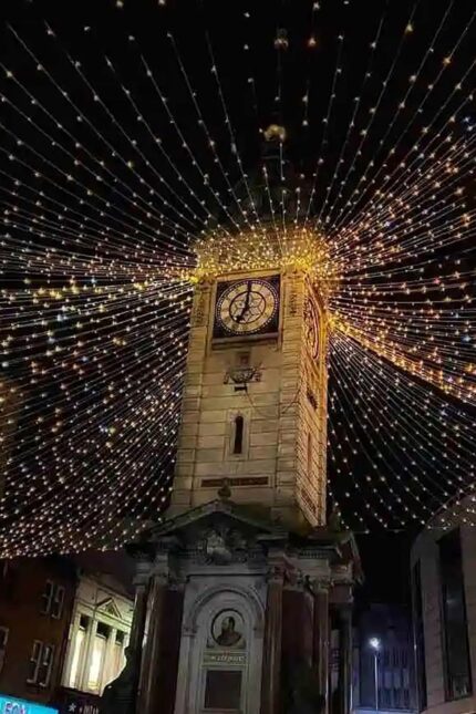 Historic stone clock tower illuminated with festive Christmas fairy lights canopy at night.