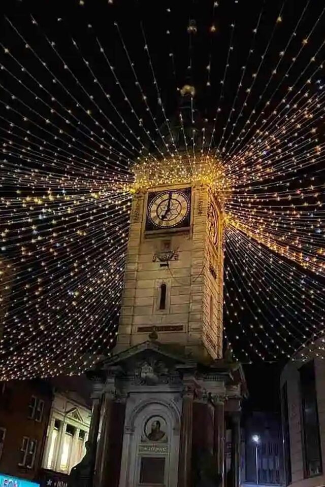 Historic stone clock tower illuminated with festive Christmas fairy lights canopy at night.