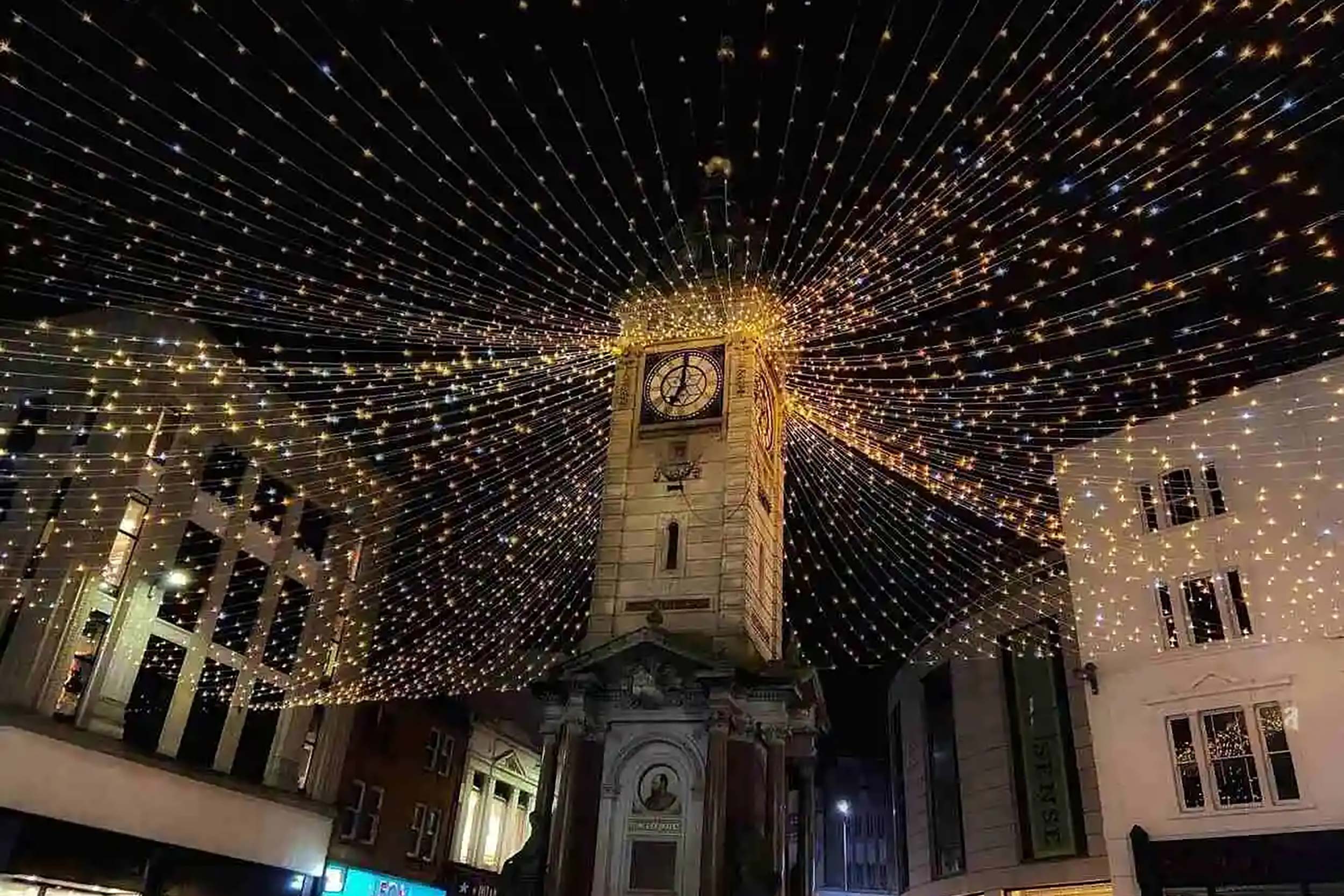 Historic stone clock tower illuminated with festive Christmas fairy lights canopy at night.