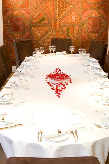 Elegant private dining table set with white linen and red petals under a tapestry wall.