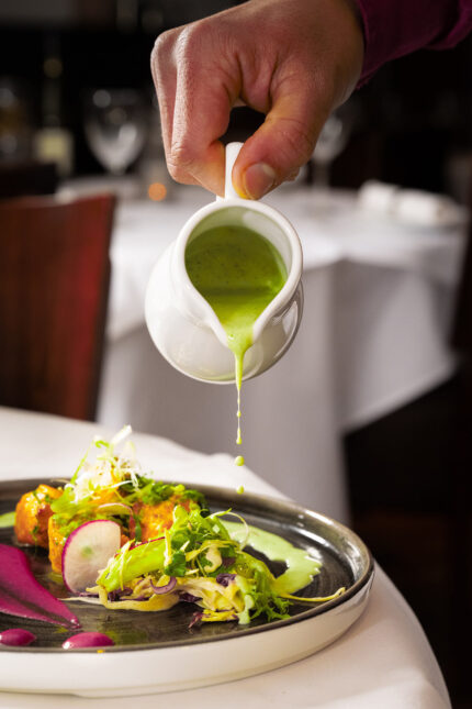 Pouring green mint sauce over an artistically plated gourmet Indian dish.