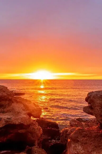 Vibrant golden sunset over the calm ocean horizon viewed from rocky coastal cliffs.